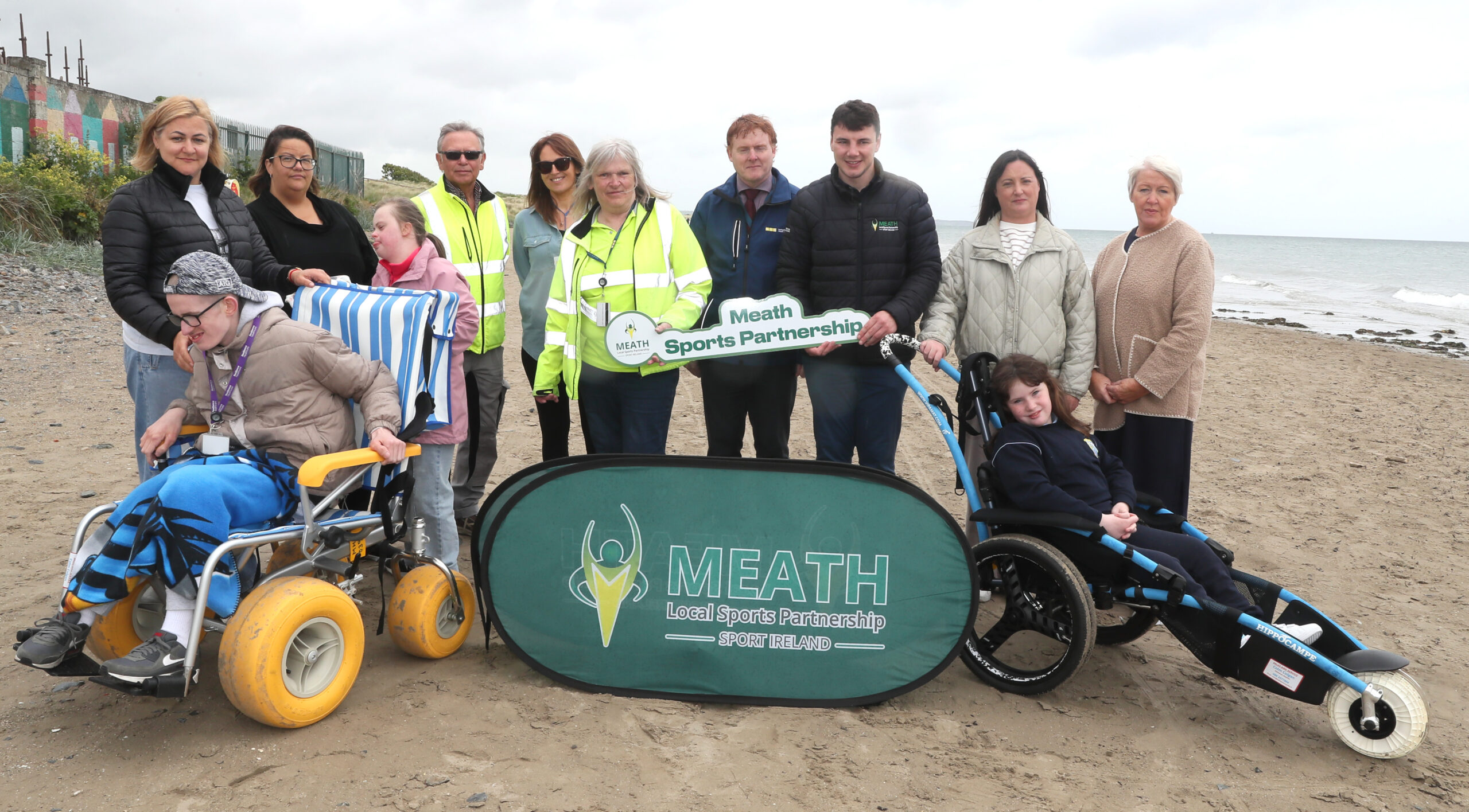 Beach Wheelchairs 5 Group of people standing on a beach with two people in beach wheelchairs