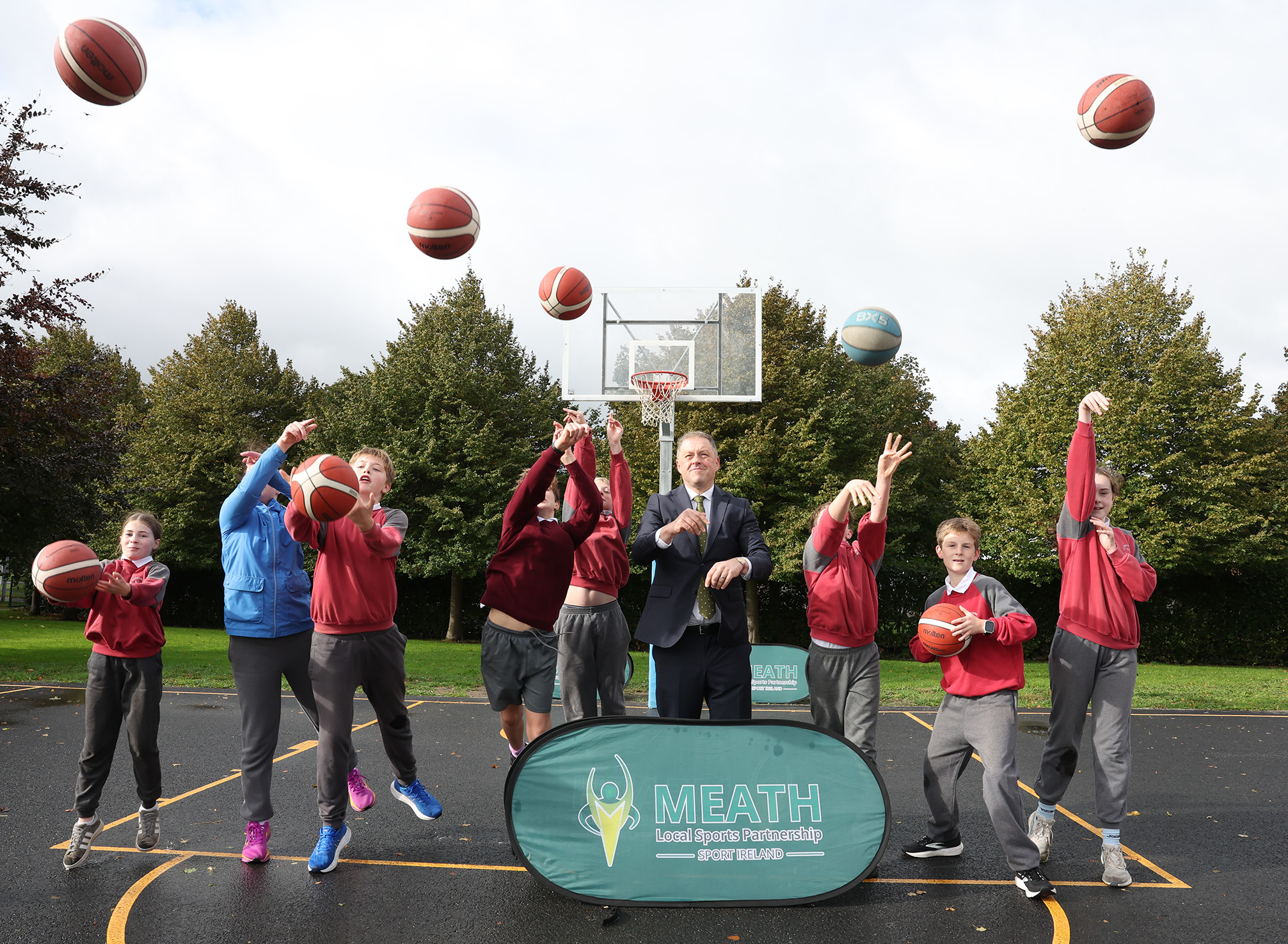 Minister Thomas Byrne T.D. with a group of schoolchildren throwing basketballs up in the air on the new 3x3 Basketball Court in Dunshaughlin.