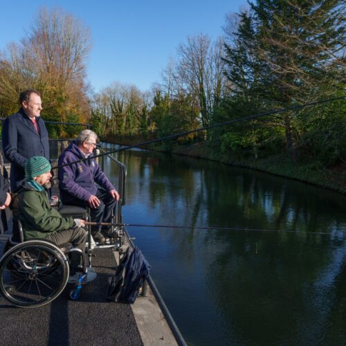 Four people stand and sit on an accessible fishing platform beside a calm canal on a sunny day. Two individuals using wheelchairs are seated at the water’s edge holding fishing rods, while two others stand behind them. Trees with autumn foliage line the opposite bank under a bright blue sky.