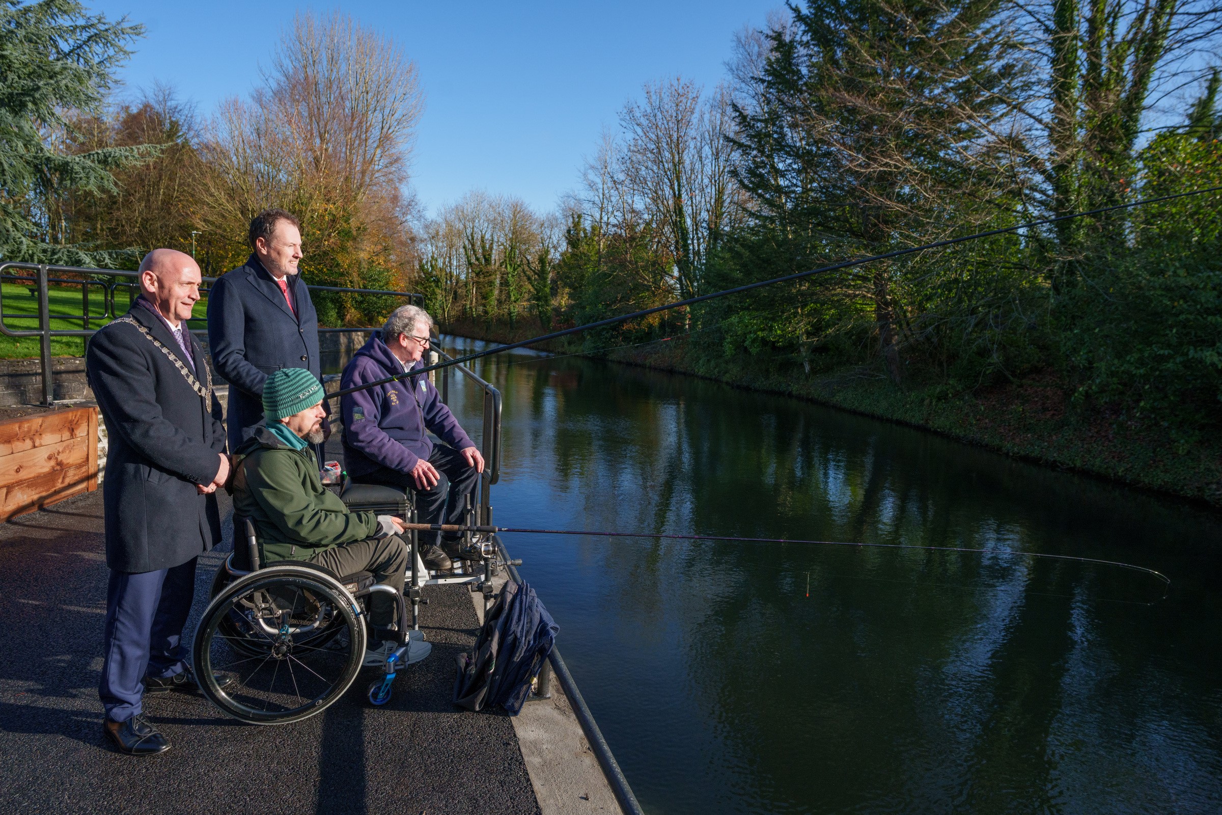 Four people stand and sit on an accessible fishing platform beside a calm canal on a sunny day. Two individuals using wheelchairs are seated at the water’s edge holding fishing rods, while two others stand behind them. Trees with autumn foliage line the opposite bank under a bright blue sky.